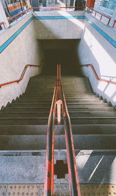 A downward view of an indoor staircase descending from a well-lit area at the top to a darker space at the bottom, with metal handrails painted orange on both sides. The staircase is made of dark stone or concrete steps with visible textures, and the walls on either side are light-colored, possibly plastered. The upper level features a corridor with a glass railing and bright ambient lighting, suggesting a modern setting such as a train station or public building. This image captures the initial portion of a home relocation or furniture transport process, where items may be carried down or up these stairs during a house move. The presence of the handrails and the lighting indicate the staircase's safety features, and the space at the top appears large enough to accommodate moving equipment or personnel preparing for a furniture loading or unloading operation, aligning with professional house removal services like those provided by Man with Van Anerley on their removals page.