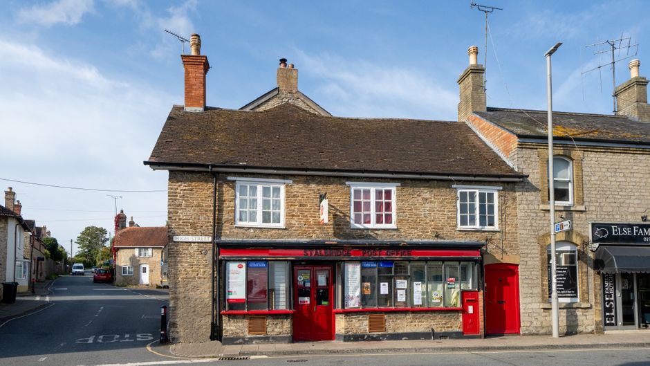A two-storey brick building located on Penge High Street in Anerley, with a red postal service window and door on the ground floor, and three white-framed windows on the upper floor. The building is a